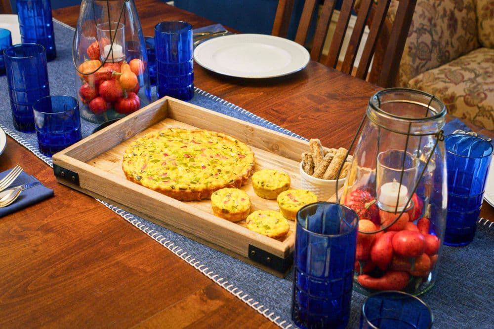 A wooden serving tray holds a frittata and small pastries, surrounded by blue glasses and fruit centerpieces on a dining table.