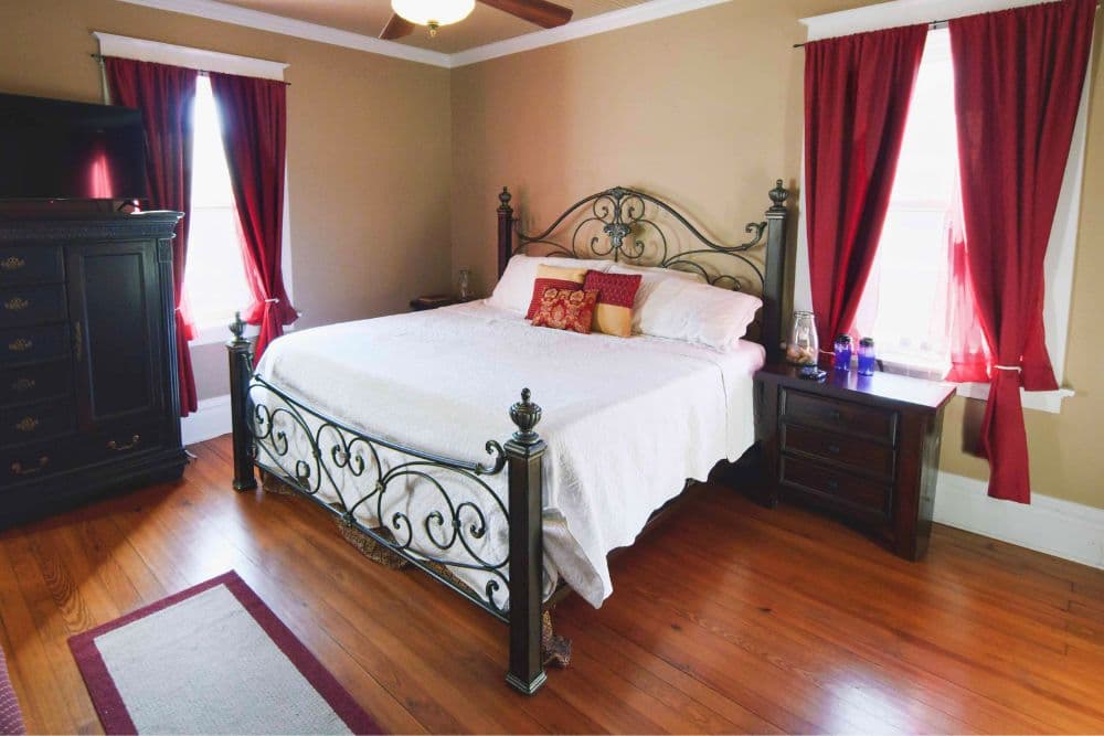A cozy bedroom featuring a wrought iron bed with white bedding, red curtains, and dark wood furniture.